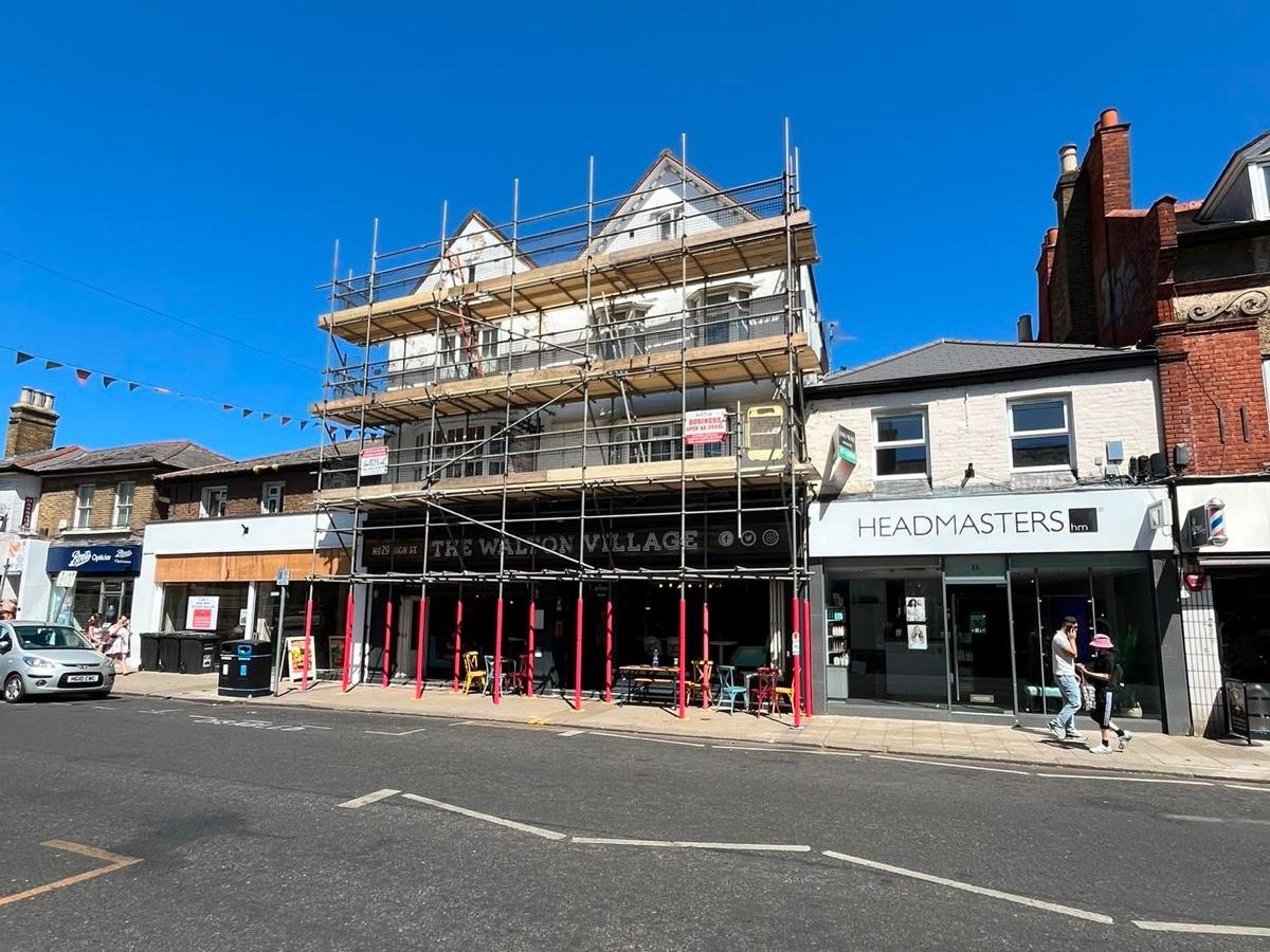 BSL scaffold across the full frontage of The Walton Village pub on Walton-on-Thames High Street, padded standards at pavement level and the pub open beneath